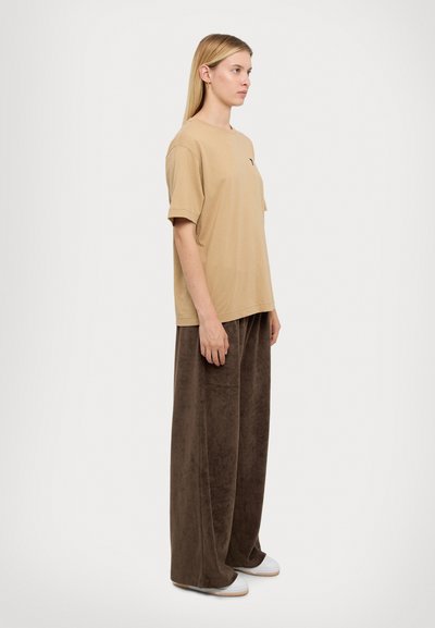 Beige Baumwoll-T-Shirt mit kurzen Ärmeln und einer Taschenöffnung, kombiniert mit braunen Velours-Hosen im weiten Schnitt und weißen Slippern.