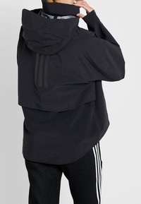 Personne ajustant la capuche d'une veste noire avec des bandes réfléchissantes verticales à l'arrière, portant un pantalon noir avec des bandes blanches sur les côtés.