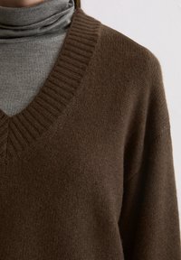 Bruine gebreide trui met een diepe V-hals en ribbel details, gelaagd over een grijze coltrui, met een zachte textuur en een casual pasvorm.