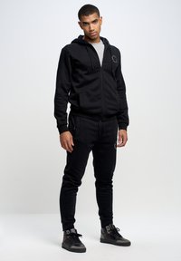 Schwarzer Reißverschluss-Hoodie mit grauem Innenfutter, kombiniert mit schwarzen Jogginghosen. Das Material wirkt weich, mit sichtbaren Taschen und strukturierten Bündchen. Schwarze Sneakers.