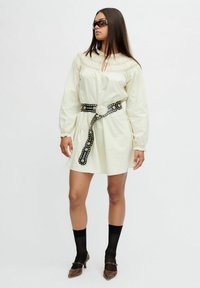 Vrouw in een crèmekleurige lange jurk met lange mouwen, een zwarte studded riem, zwarte sheer sokken, bruine hakken en donkere zonnebril, staand tegen een witte achtergrond.