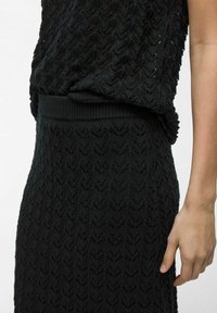 Robe tricotée noire présentant un motif zigzag texturé, une taille côtelée et une coupe décontractée. Le tissu semble doux et léger.