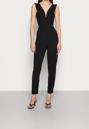 Zwarte jumpsuit met een diepe V-hals en ruches langs de schouders, aansluitend ontwerp en tapse pijpen. Gecombineerd met zwarte sandalen.