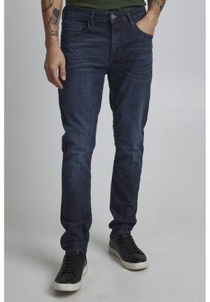 Jean en denim bleu foncé avec une coupe slim, comportant cinq poches et des détails de couture subtils, associés à des baskets noires basses.