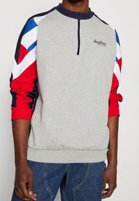 Maglione in pile grigio con mezza cerniera, caratterizzato da maniche con motivo rosso, bianco e blu e colletto e polsini a coste.