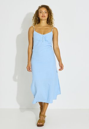 Femme aux cheveux bouclés portant une robe midi sans manches bleu clair et des sandales compensées marron, marchant vers l'avant sur un fond blanc uni.