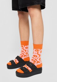 Black platform sandals with two wide straps. Orange socks featuring pink floral patterns. Worn with black shorts.