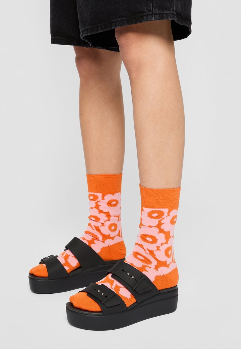 Black platform sandals with two wide straps. Orange socks featuring pink floral patterns. Worn with black shorts.