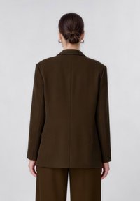 Femme aux cheveux foncés attachés en chignon, portant un blazer ajusté vert olive et un pantalon assorti, debout devant un fond clair uni.