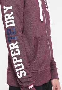 Sudadera con cremallera en color burdeos, hecha de tejido texturizado, con letras blancas "SUPER DRY" en la manga y una etiqueta de logo en el dobladillo.