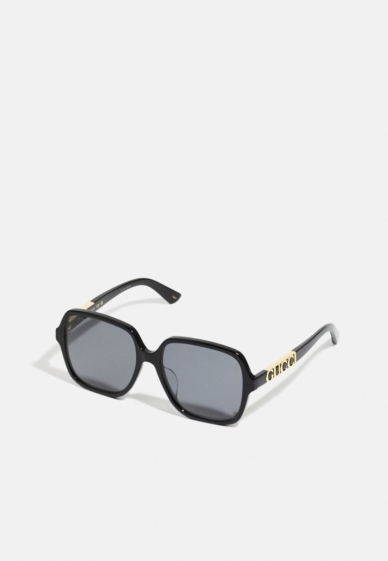 Lunettes de soleil noires avec une monture carrée, verres gris, finition polie et branches rehaussées d'or portant le nom de la marque "GUCCI."