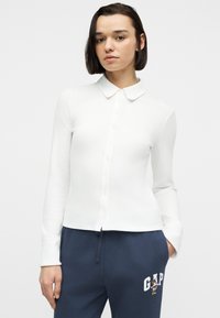 Γυναίκα με κοντά σκούρα μαλλιά φοράει λευκό πουκάμισο με κουμπιά και μακρυμάνικο, μπλε σκούρο παντελόνι φόρμας με λογότυπο GAP και γραφικό της Minnie Mouse.