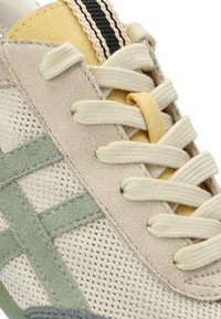 Zapatillas beige que presentan materiales de malla y ante, acentos en verde y amarillo, cordones planos y una suela texturizada con un diseño en capas.