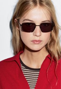 Czarne prostokątne okulary przeciwsłoneczne z ciemnymi soczewkami, z cienkimi metalowymi ramkami. Temat nosi czerwoną koszulę na czarno-białą pasiastą bluzkę.