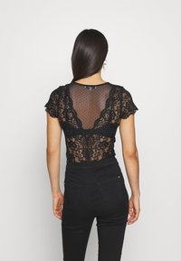 Černý krajkový body se krátkým rukávem, s průhledným puntíkovaným zadním dílem a květinovými vzory. V kombinaci s černými kalhotami s vysokým pasem.