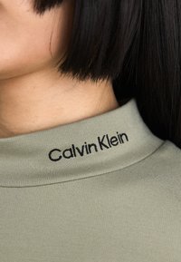Olive green fitted top with a high collar, featuring black embroidered text "Calvin Klein" at the neckline. Smooth, textured fabric.
