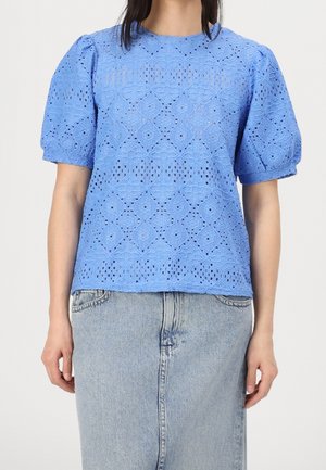Jasnoniebieska bluzka z krótkim rękawem, charakteryzująca się teksturowanym wzorem w ażurowe kółka oraz bufiastymi rękawami. Połączona z długą spódnicą dżinsową z wysokim stanem.