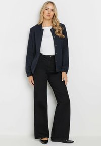 Giacca trapuntata blu navy con colletto rotondo, bottoni a pressione, abbinata a una camicia bianca e pantaloni larghi neri. Texture liscia, design informale.