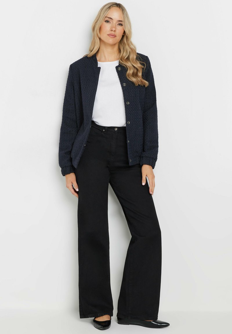 Giacca trapuntata blu navy con colletto rotondo, bottoni a pressione, abbinata a una camicia bianca e pantaloni larghi neri. Texture liscia, design informale.