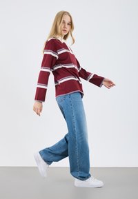 Chemise à manches longues bordeaux avec des rayures blanches et bleues, associée à un jean large bleu clair et des baskets blanches, sur un fond neutre.