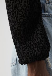 Svart leopardmønstret jakkeerm med strammet elastikkant. Materialet ser lett og gjennomsiktig ut, med en teksturert overflate. Denim bakgrunn.
