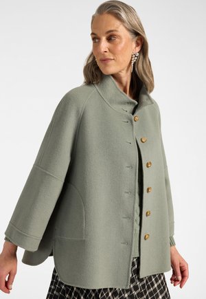 Donna con capelli grigi che indossa un cappotto di lana verde salvia abbottonato e orecchini pendenti di perle, che guarda di lato su uno sfondo bianco.