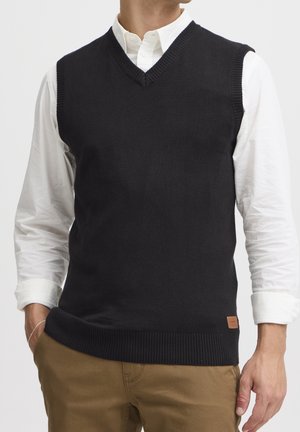 Homme portant un pull sans manches en tricot noir à col en V par-dessus une chemise blanche à manches longues et un pantalon marron, une main dans la poche.