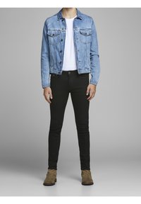 Persona con chaqueta de mezclilla azul, camiseta blanca, pantalones ajustados negros y botas de ante marrón, de pie contra un fondo gris.