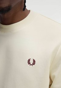 Felpa di colore crema con colletto a costine e un piccolo logo ricamato color bordeaux sul davanti. Tessuto morbido e vestibilità rilassata.