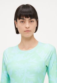 Long-sleeve, light teal rash guard with a floral pattern. The material is smooth and stretchy, featuring a crew neck and raglan sleeves.