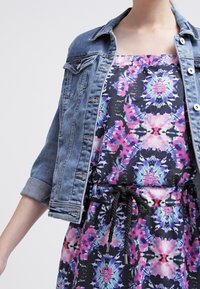 Veste en denim avec accents usés sur une robe colorée à motifs comportant des designs abstraits en rose, bleu et noir, avec une taille à cordon.