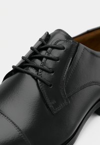 Zapato de vestir de cuero negro con acabado brillante, puntera redonda y diseño tradicional con cordones. Presenta costuras sutiles y una textura suave.