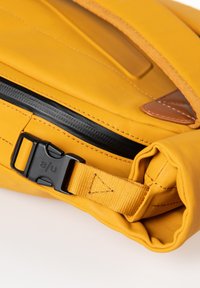 Borsa in tessuto giallo con una cerniera nera, un accento in pelle marrone e una tracolla nera con fibbia. Il materiale sembra liscio con dettagli di cucitura visibili.