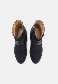 Stivaletti alla caviglia in suede blu navy con un design liscio e curvo, dotati di dettagli in pelle e una cerniera laterale. Suola piatta e punta arrotondata.