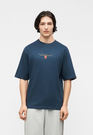 Młody mężczyzna o średniej długości ciemnych włosach, ubrany w granatowy t-shirt z grafiką i jasnoszare spodnie, stojący na jednolitym tle.