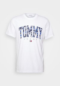 Bílý tričko s krátkým rukávem se znakem "TOMMY" v modré barvě s designem, "JEANS" v černé barvě pod ním a malou logem jako akcent. Materiál: bavlna.