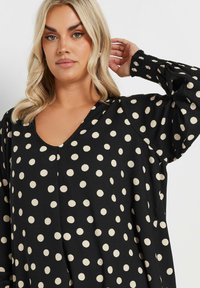 Svart blus i tyg med stora, krämfärgade prickar. Designen har en V-ringning och voluminösa ärmar med smockade manschetter.