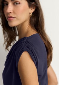 Tengerészkék felső rövid, fodros ujjakkal, sima anyagból, laza fazonban, amelyet egy hosszú, hullámos barna hajú nő visel. Arany karika fülbevalók.