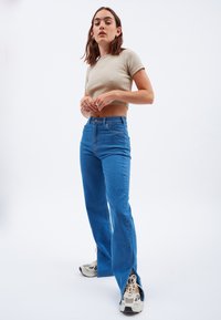 Top corto de color beige claro con mangas cortas, combinado con unos jeans acampanados azules de cintura alta. La modelo lleva zapatillas grises y blancas.