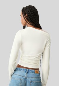 Hvid langærmet top med rund halsudskæring, tætsiddende design og draperet detalje i siden, sammen med blå højtaljede jeans.