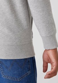 Szary sweatshirt z prążkowymi mankietami i dołem, prezentujący miękką fakturę. Nosi się go z niebieskimi dżinsami wyróżniającymi się charakterystycznym wzorem przeszyć.