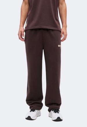 Άνδρας που φορά σκούρες καφέ φόρμες και αντίστοιχο πουκάμισο, όρθιος, με λευκά αθλητικά παπούτσια σε μονόχρωμο φόντο.