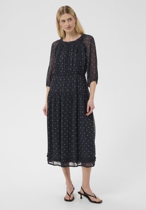 Femme debout portant une robe midi noire transparente à pois blancs et manches trois-quarts, associée à des sandales noires à talons.