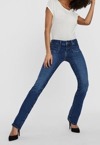 Blå denimjeans med rak skärning, medelhög midja, fem fickor och svaga slitningar, bärs tillsammans med en vit t-shirt och svarta klackar.