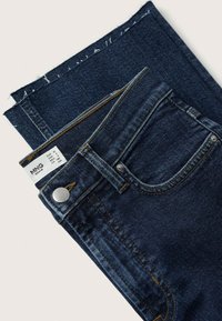 Jeans de mezclilla azul oscuro doblados, con el bolsillo frontal visible, el lazo del cinturón, el botón y la etiqueta de tamaño que muestra las tallas europeas y estadounidenses.