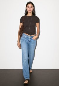 Dunkelbraunes, tailliertes T-Shirt, hellblaue weite Jeans, schwarze Sandalen und eine strukturierte braune Umhängetasche. Einfaches silbernes Halskettchen mit Anhänger.