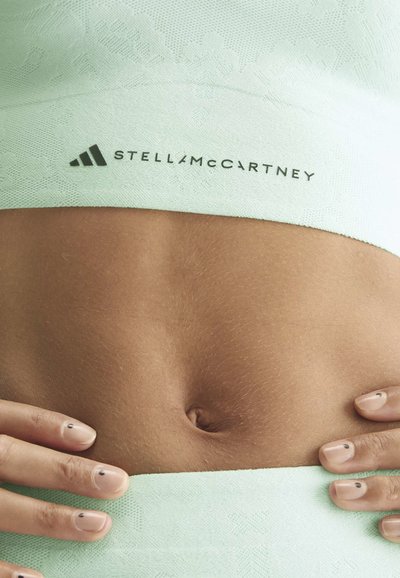 Nahaufnahme vom nackten Bauch, der ein hellgrünes, strukturiertes Gewebe mit Stella McCartney Logo trägt, und Hände mit einfacher schwarzer Punkt-Nagelkunst.