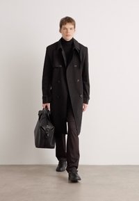 Sort dobbeltradet trench coat i uldblanding, parret med en sort turtleneck og mørke bukser. Bærer en stor lædertaske. Glat tekstur.
