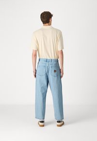 Camiseta beige claro de manga corta, combinada con unos vaqueros de tela vaquera azul claro de corte holgado, que cuentan con bolsillos traseros y una etiqueta de marca.