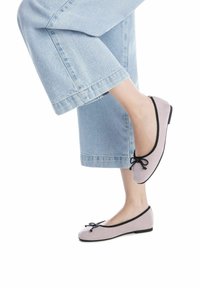 Bailarinas de ante rosa claro con ribete negro y detalle de lazo, combinadas con unos jeans de denim cortos. Los zapatos presentan un perfil bajo y una suela flexible.